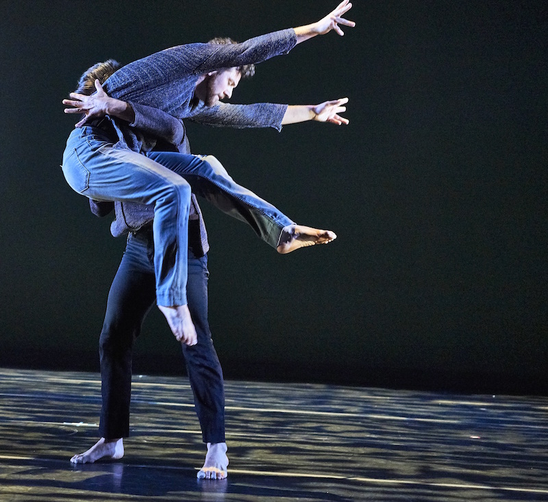 One man in jeans hovers in the air while another catches him by his waist. 